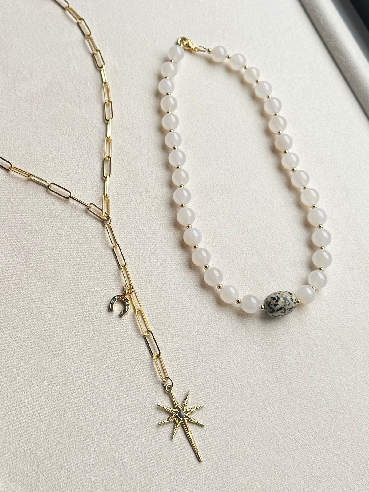 Two necklaces displayed on a light beige textured surface: one is a gold-tone chain with paperclip links, a small crescent horseshoe charm, and a starburst charm; the other is a white agate beaded necklace, spaced with gold spacer beads and a single dalmation jasper stone accent.