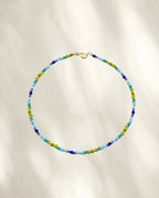 A delicate beaded necklace with glass seed beads in light blue, medium blue, and lime green, accented by a gold clasp, displayed on a textured cream fabric background with soft shadows.