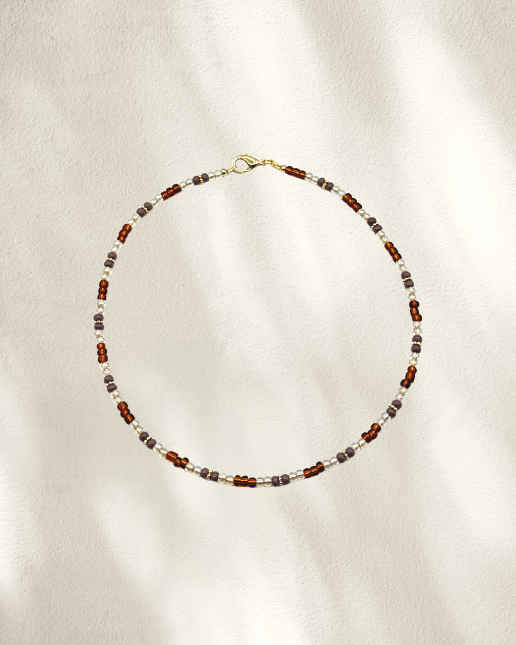 A delicate beaded necklace with alternating brown, clear/silver and gray/purple glass beads, accented by small gold spacer beads and a gold clasp, displayed on a textured cream fabric with soft shadows.