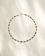 A delicate beaded necklace with alternating brown, clear/silver and gray/purple glass beads, accented by small gold spacer beads and a gold clasp, displayed on a textured cream fabric with soft shadows.