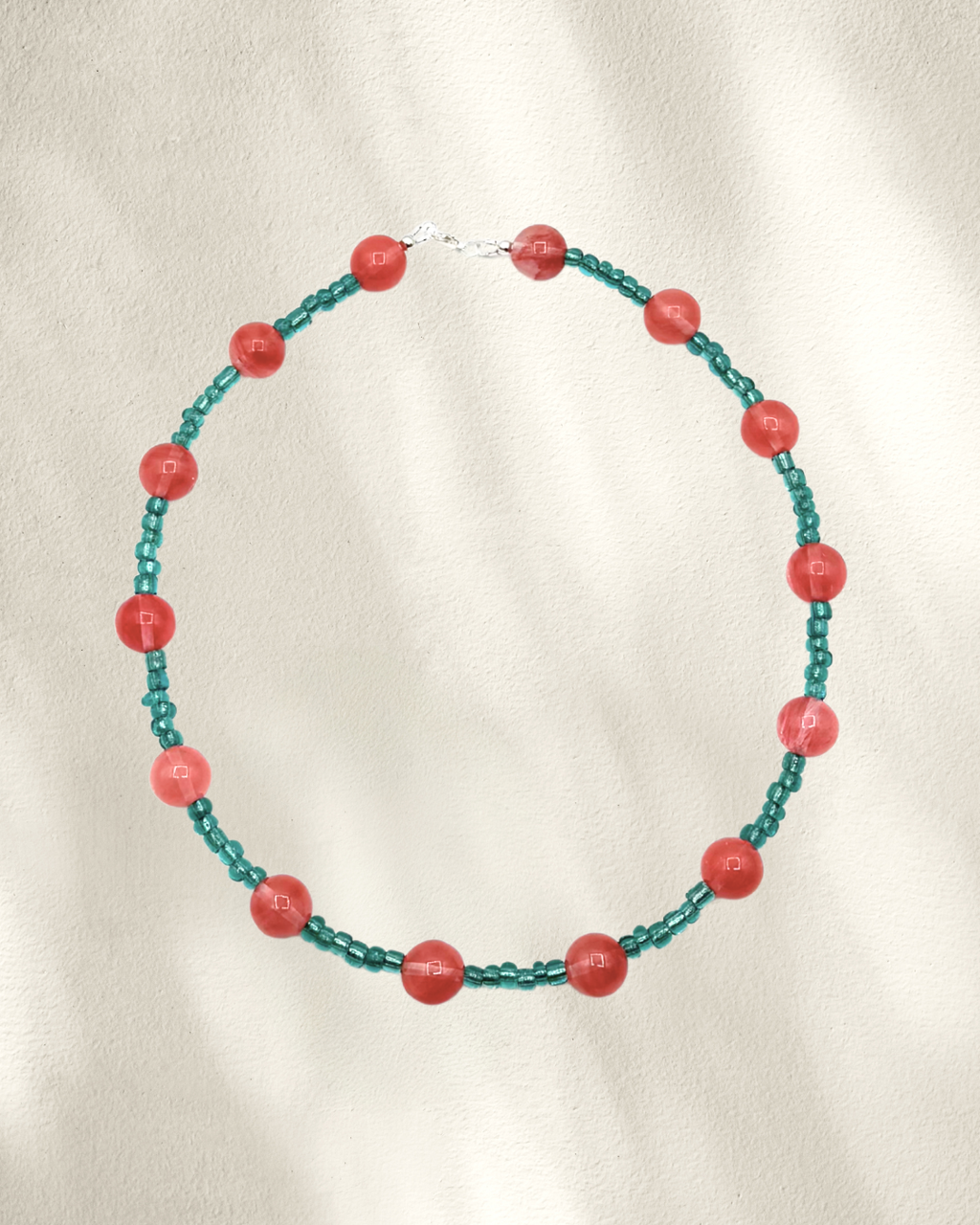A beaded necklace featuring alternating translucent teal green glass beads and larger coral-red cherry quartz round beads, arranged in a circle on a textured off-white fabric background.