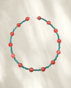 A beaded necklace featuring alternating translucent teal green glass beads and larger coral-red cherry quartz round beads, arranged in a circle on a textured off-white fabric background.