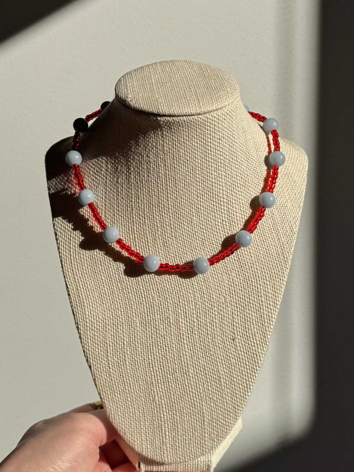 A beaded necklace with red glass seed beads and light gray/blue Angelite round beads, displayed on a beige fabric jewellery bust. A hand is visible holding the base of the bust, and the background is a warm beige wall with strong sunlight and shadows.