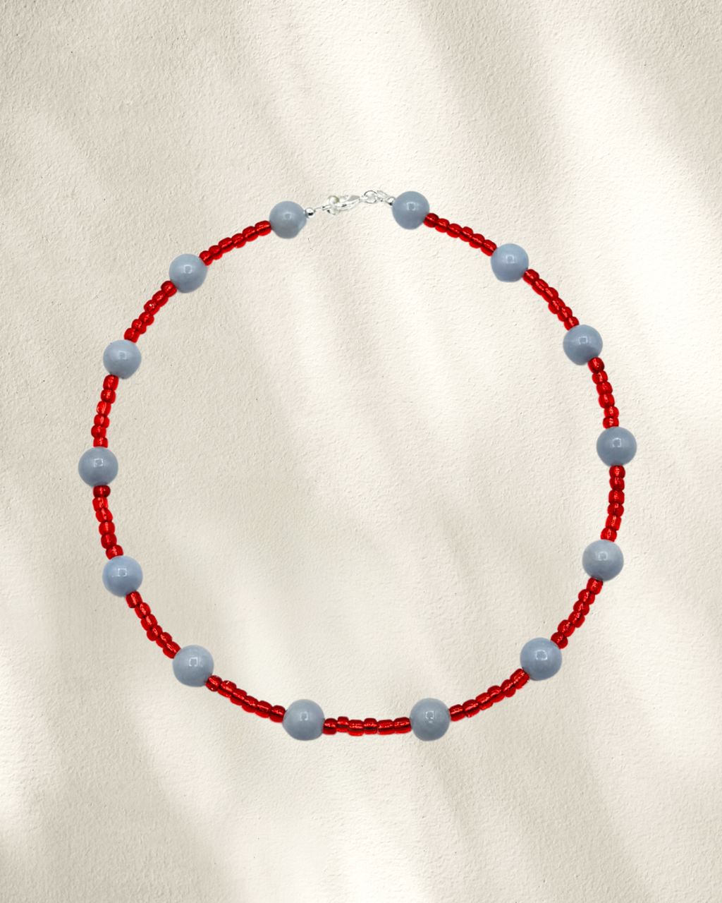 A beaded necklace featuring red glass seed beads and alternating smooth, matte light blue-gray Angelite round beads, displayed on a textured cream background.