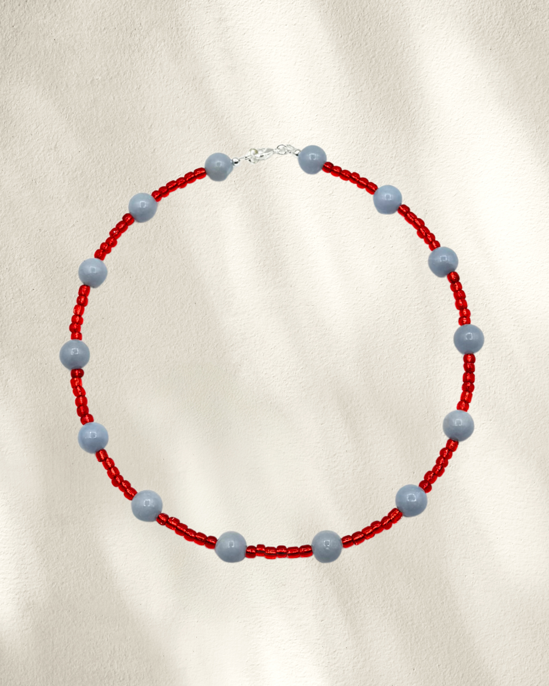 A beaded necklace featuring red glass seed beads and alternating smooth, matte light blue-gray Angelite round beads, displayed on a textured cream background.