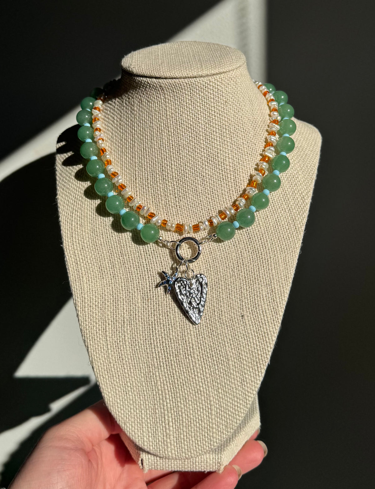 Beaded necklaces displayed on a beige fabric jewellery bust, featuring green adventurine beads, blue glass beads orange glass seed beads and freshwater pearls with a silver molten heart and starfish pendant. A hand is holding the base of the bust against a dark background.