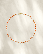 A delicate beaded necklace with small orange glass seed beads separated by freshwater pearls, accented with tiny gold details and a gold clasp, laid flat on a textured off-white background.