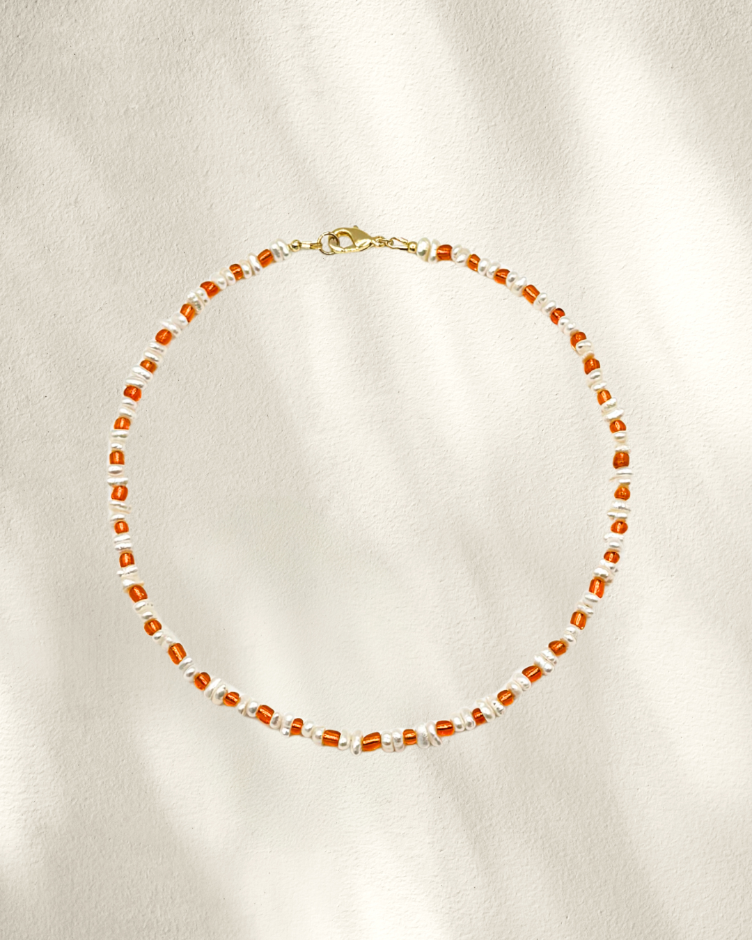 A delicate beaded necklace with small orange glass seed beads separated by freshwater pearls, accented with tiny gold details and a gold clasp, laid flat on a textured off-white background.